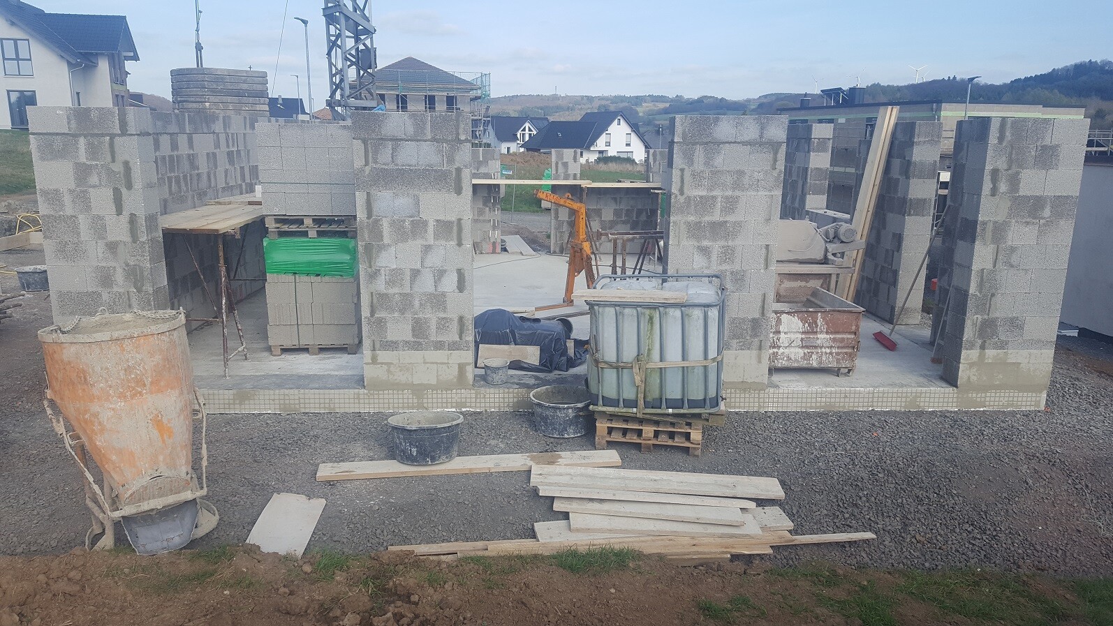 Unfertige Baustelle mit Betonblockwänden, Holzplanken, Eimern und Baumaschinen auf Kies.
