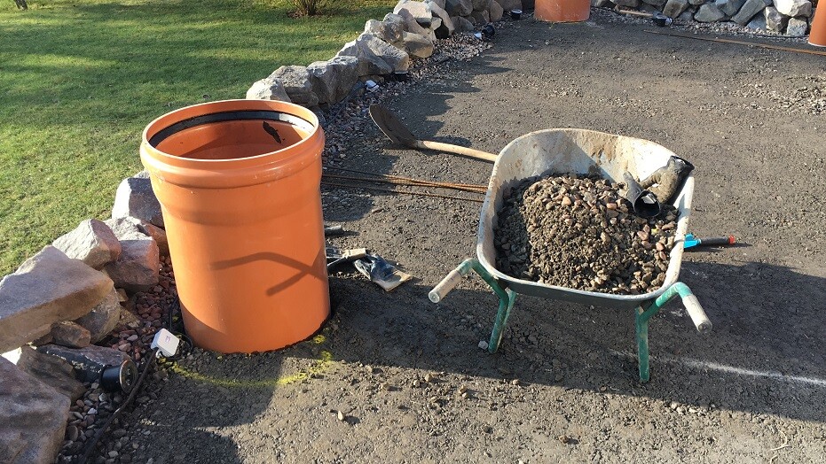 Gartenbaustelle: großes orangefarbenes PVC-Rohr neben Schubkarre voller Kies.