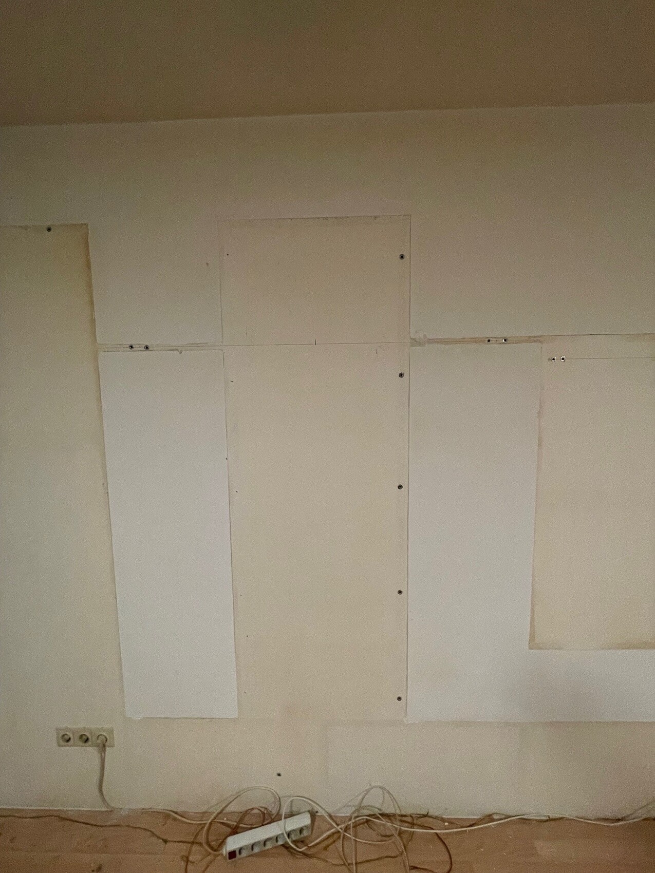 Weiße Wand mit sichtbaren Drywall-Patches, Kabelsalat und Steckdosenleiste am Boden.