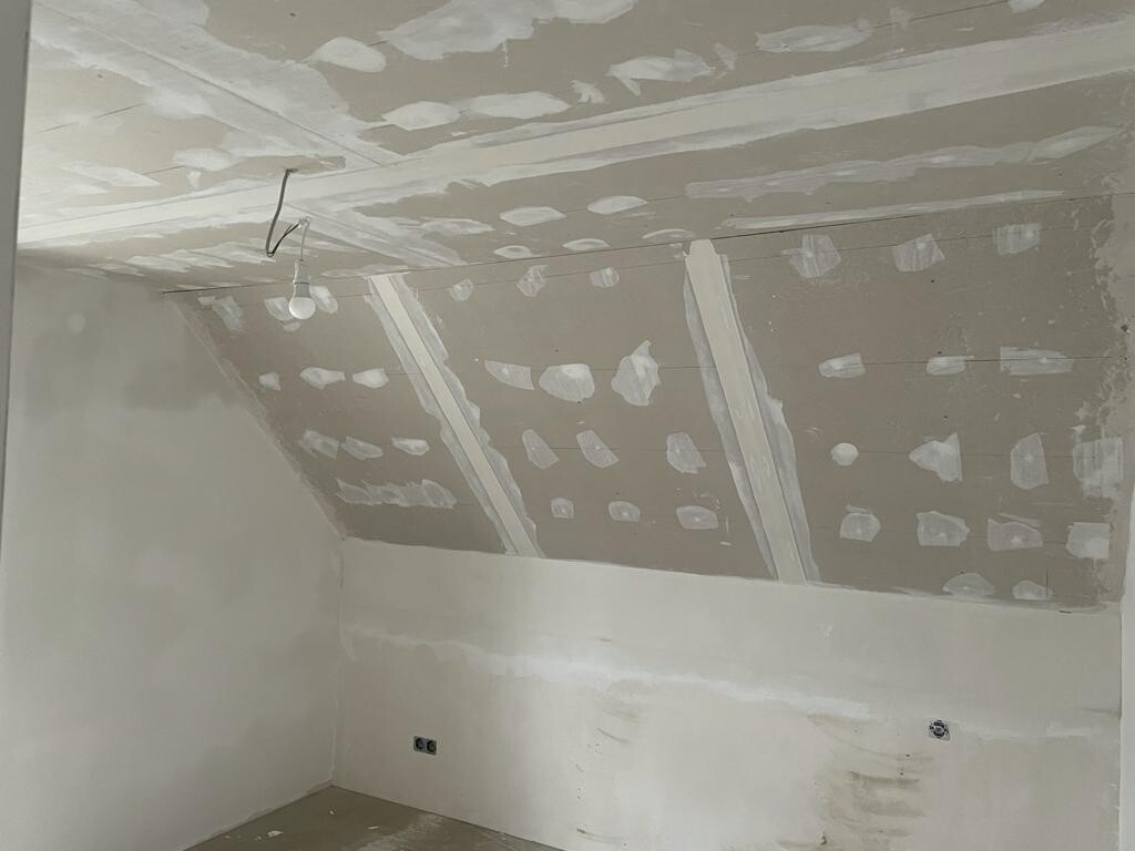 Unfertiges Zimmer mit gespachtelten Trockenbauflächen, mit einer hängenden Glühbirne und lose Kabel.