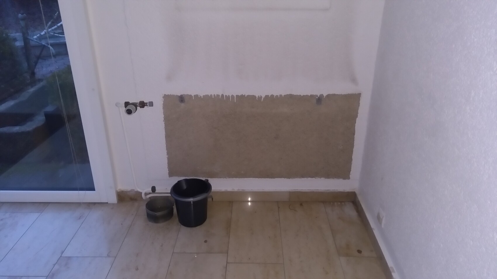 Weiße Wand mit großem braunen Putzfleck und Tropfspuren; links Wasserhahn, Eimer auf Fliesenboden.