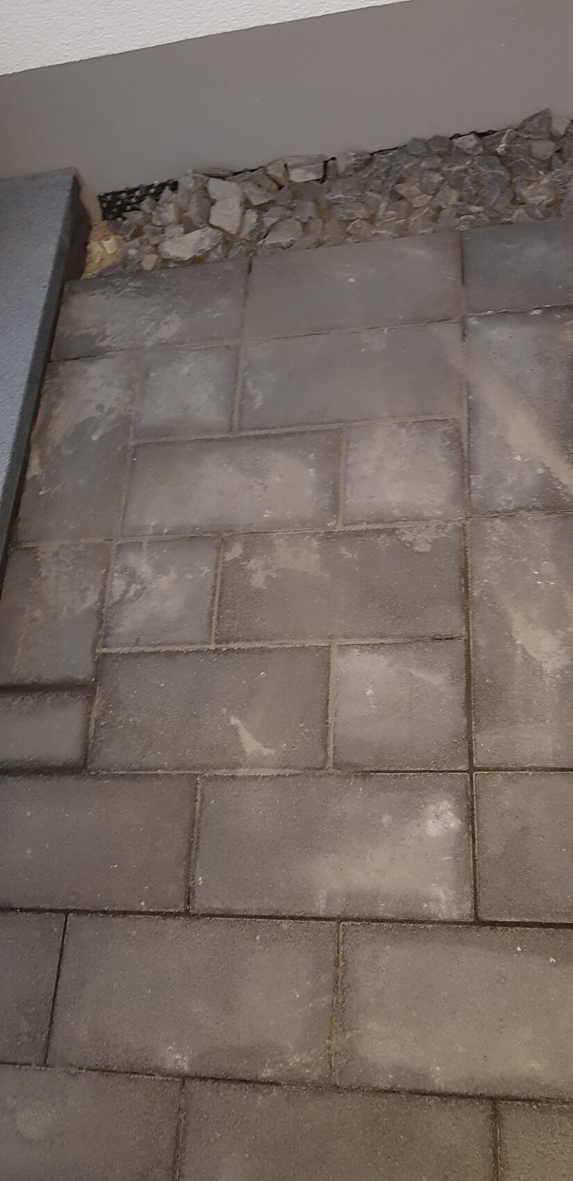 Grau gepflasterter Boden aus rechteckigen Betonplatten; am Rand liegen lose Kiesel.