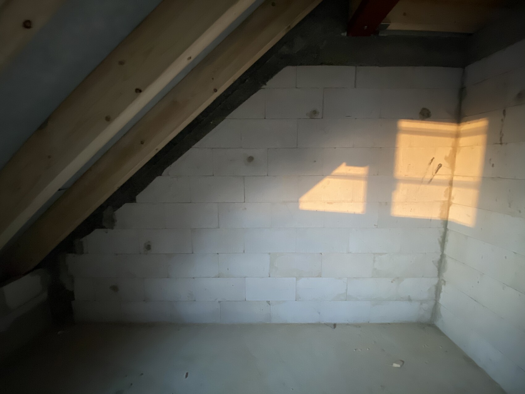 Dachboden mit Holzbalken und weißen Steinblöcken; Sonnenlicht wirft rechteckige Lichtfläche an Wand.