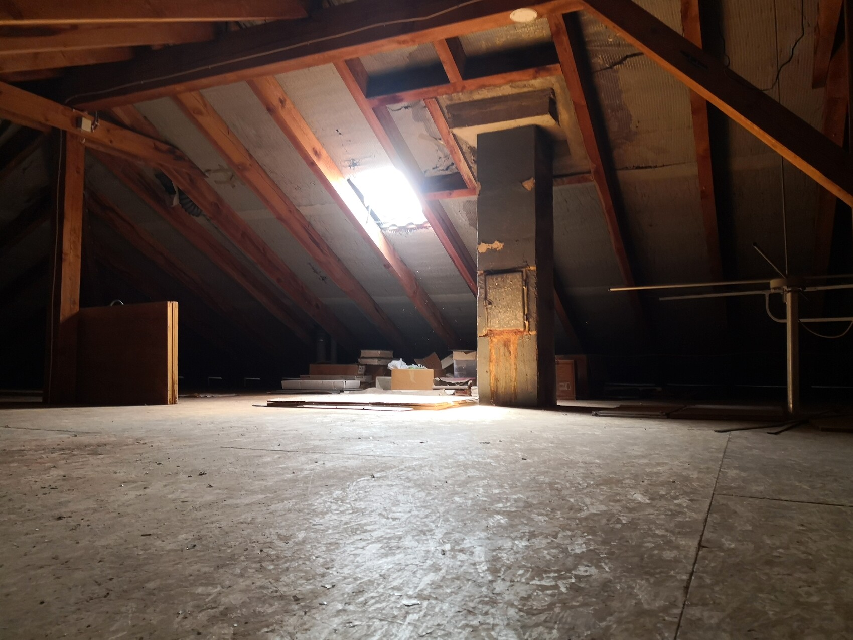 Dunkler Dachboden mit Holzbalken, zentrale Stütze, Lichtstrahl durchs Dachfenster, Kisten und Holzreste.