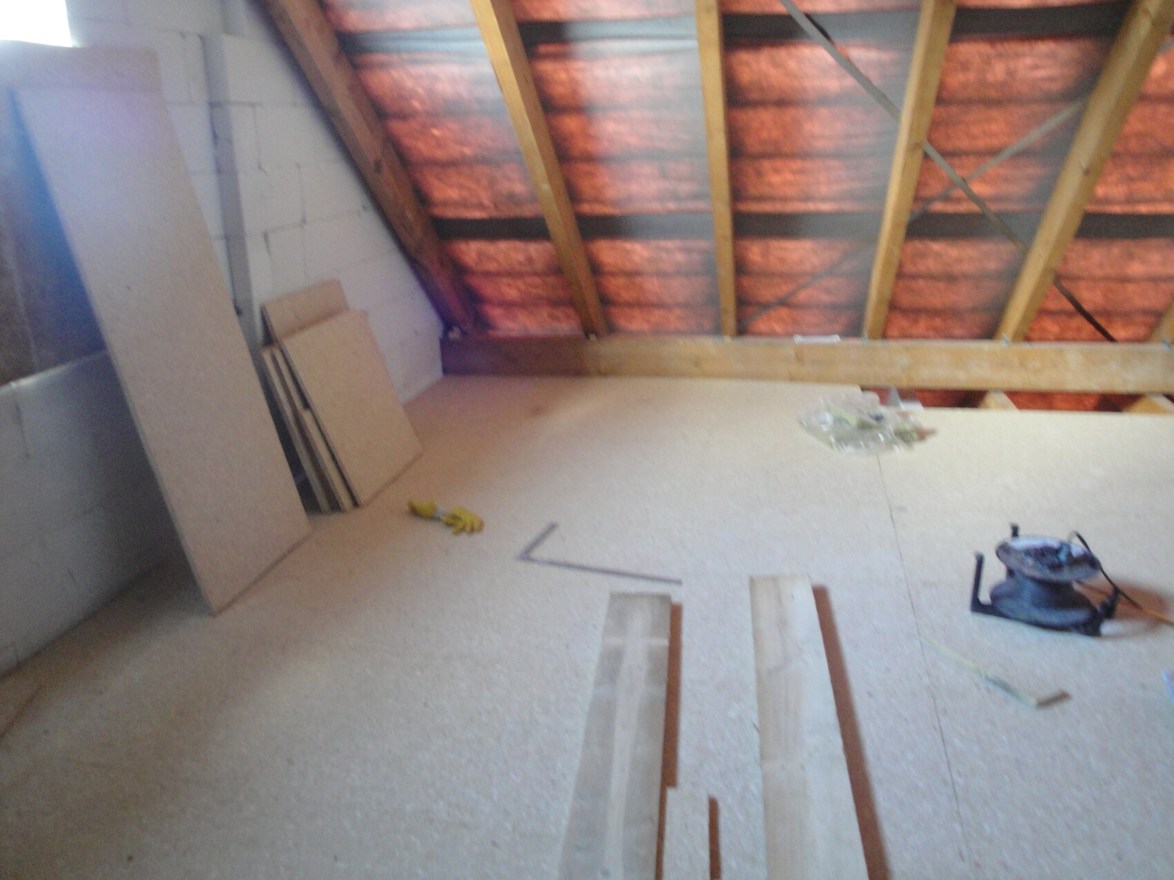 Dachboden mit sichtbarem Dachstuhl, orange Dämmung, Holzplatten und Werkzeug auf dem Boden.
