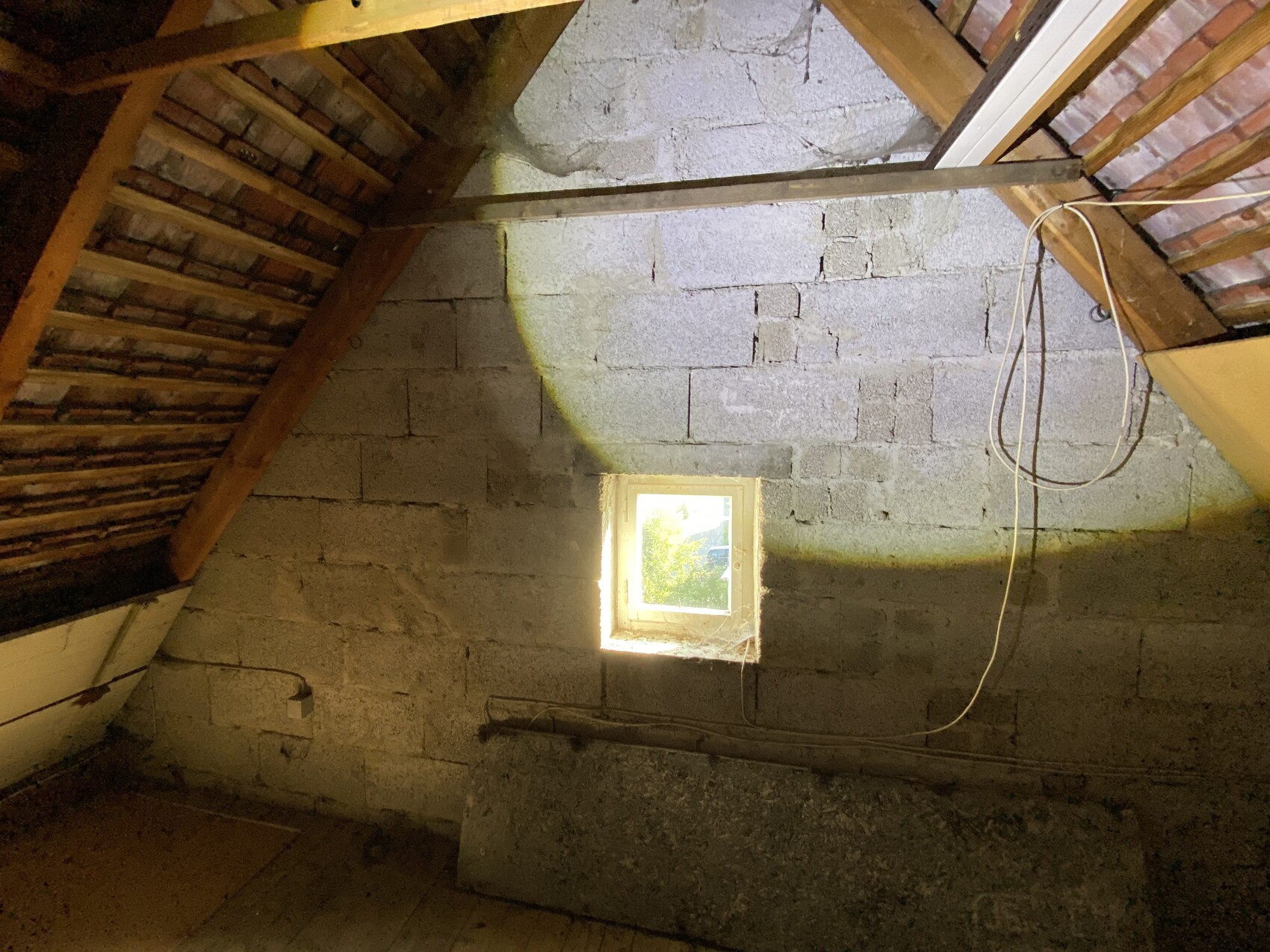 Dachboden im Rohbau mit Holzbalken, kleinem Fenster und rauen Betonblockwänden