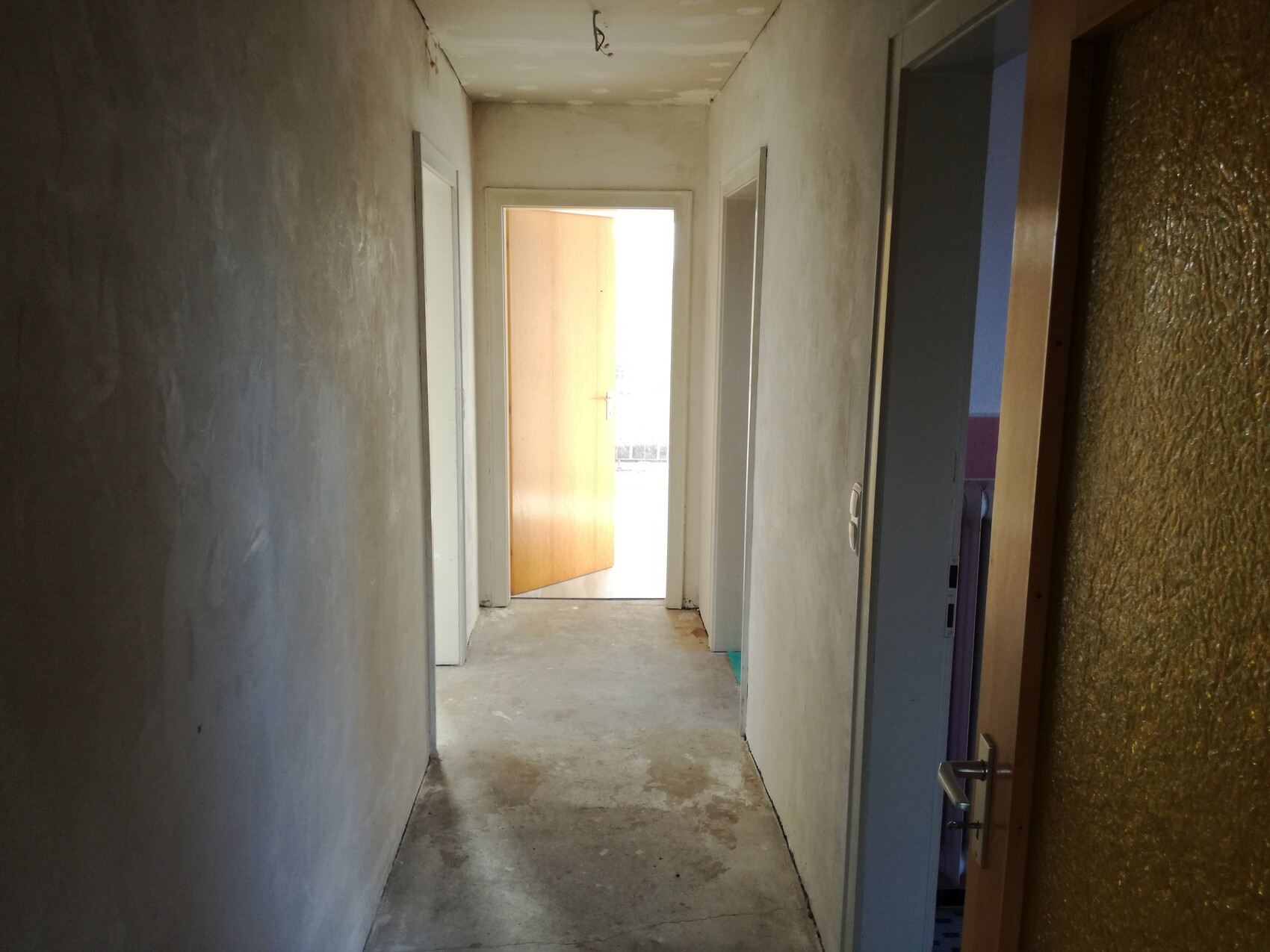 Dunkler Flur mit rauen Wänden; am Ende eine offene Holztür führt nach draußen ins Licht.