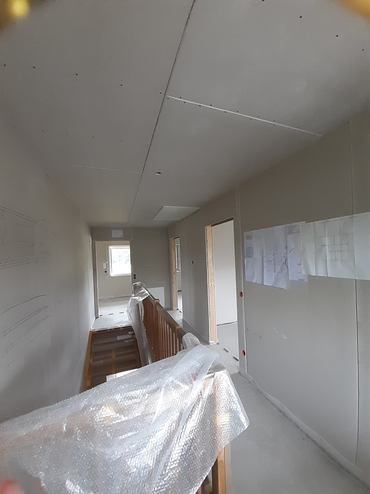 Heller Bauflur mit Treppenaufgang, Geländer mit Schutzfolie, Türen und Bauplänen an der Wand.