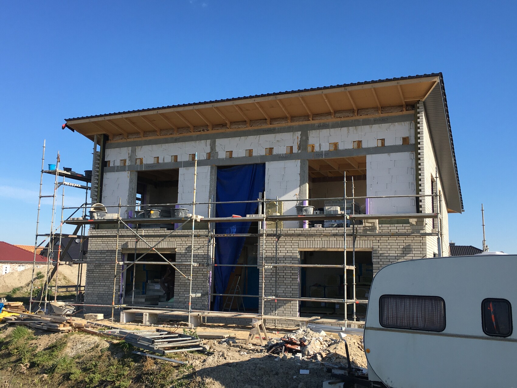 Zweistöckiges Haus im Rohbau mit Gerüst, Baumaterialien und blauer Plane; Bauwagen vorne.