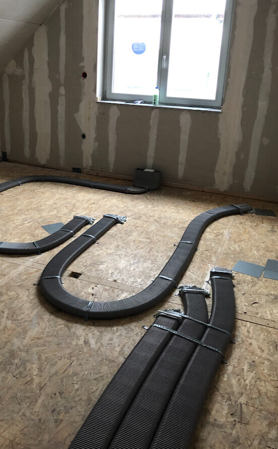 Baustelle: Schwarze flexible Rohre verlaufen über Holzboden; Fenster im Hintergrund.