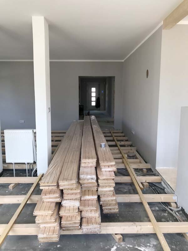 Innenraum eines Neubaus: Stapel Holzplanken auf Unterbau, graue Wände und Durchgang in Sicht.