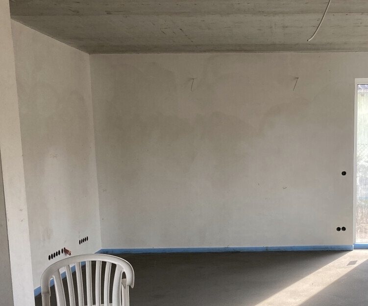 Unfertiger Innenraum mit Rohbauwänden, sichtbaren Steckdosen und blauer Bodenschutzkante.