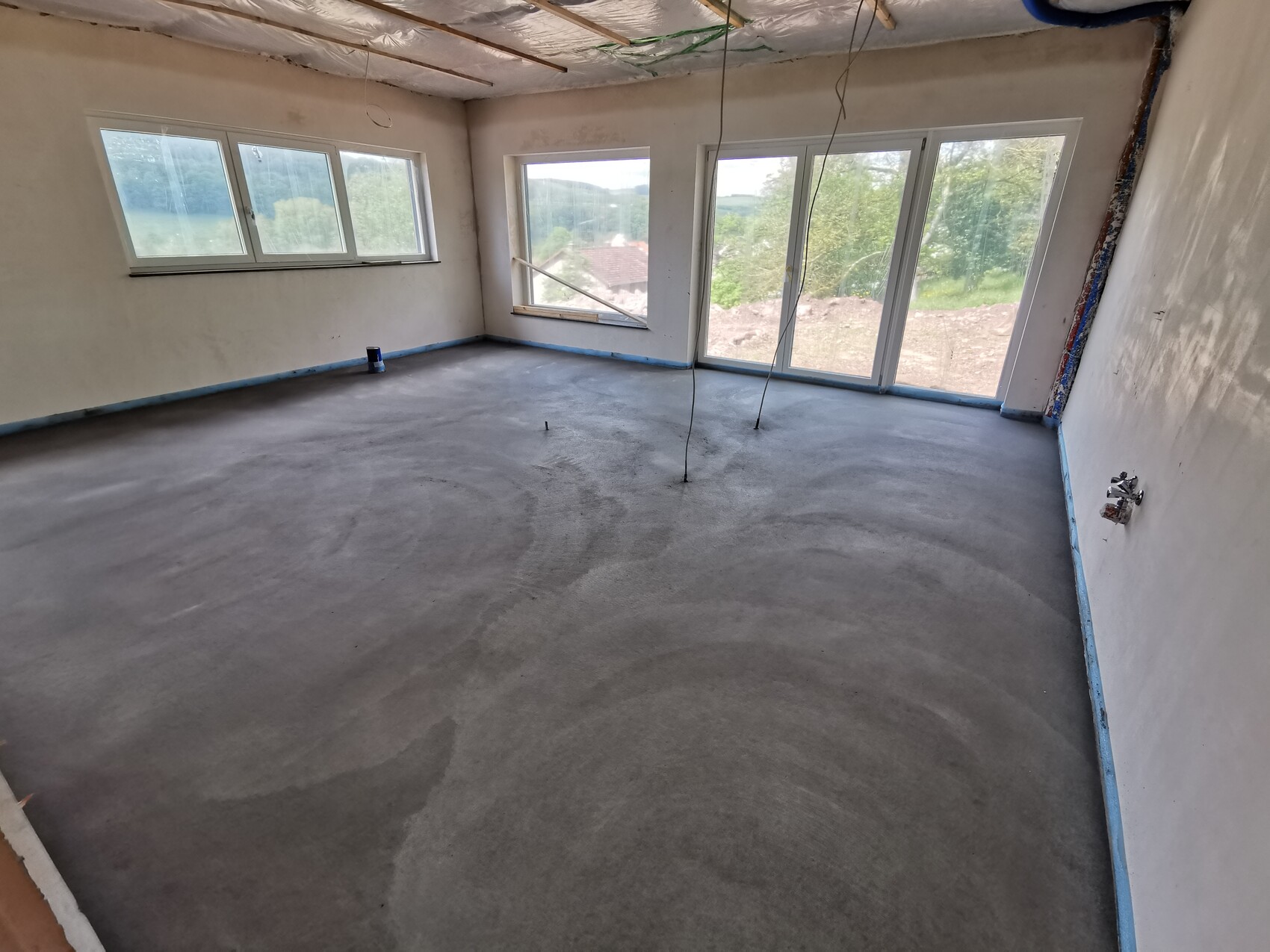 Unfertiger Wohnraum unter Bau: glatter Betonboden, lose Kabel, große Fensterfront.