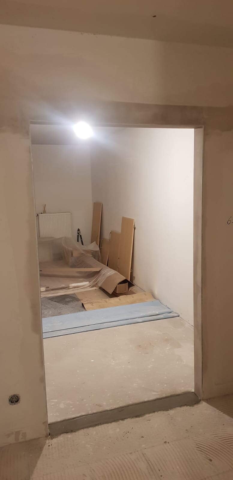 Unfertiger Innenraum einer Baustelle: Türöffnung, Holzplatten, Karton, Plastikfolie, Licht.