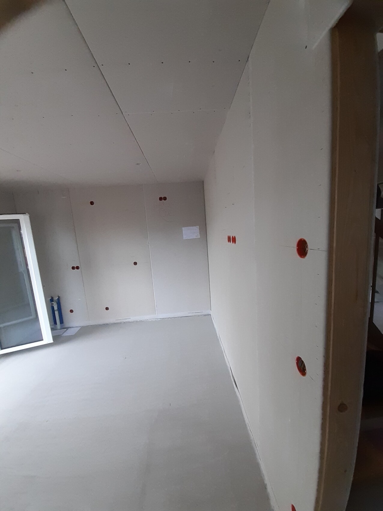 Unfertiger Innenraum mit weißen Gipskartonwänden, roten Steckdosenöffnungen und Fensterrahmen links.
