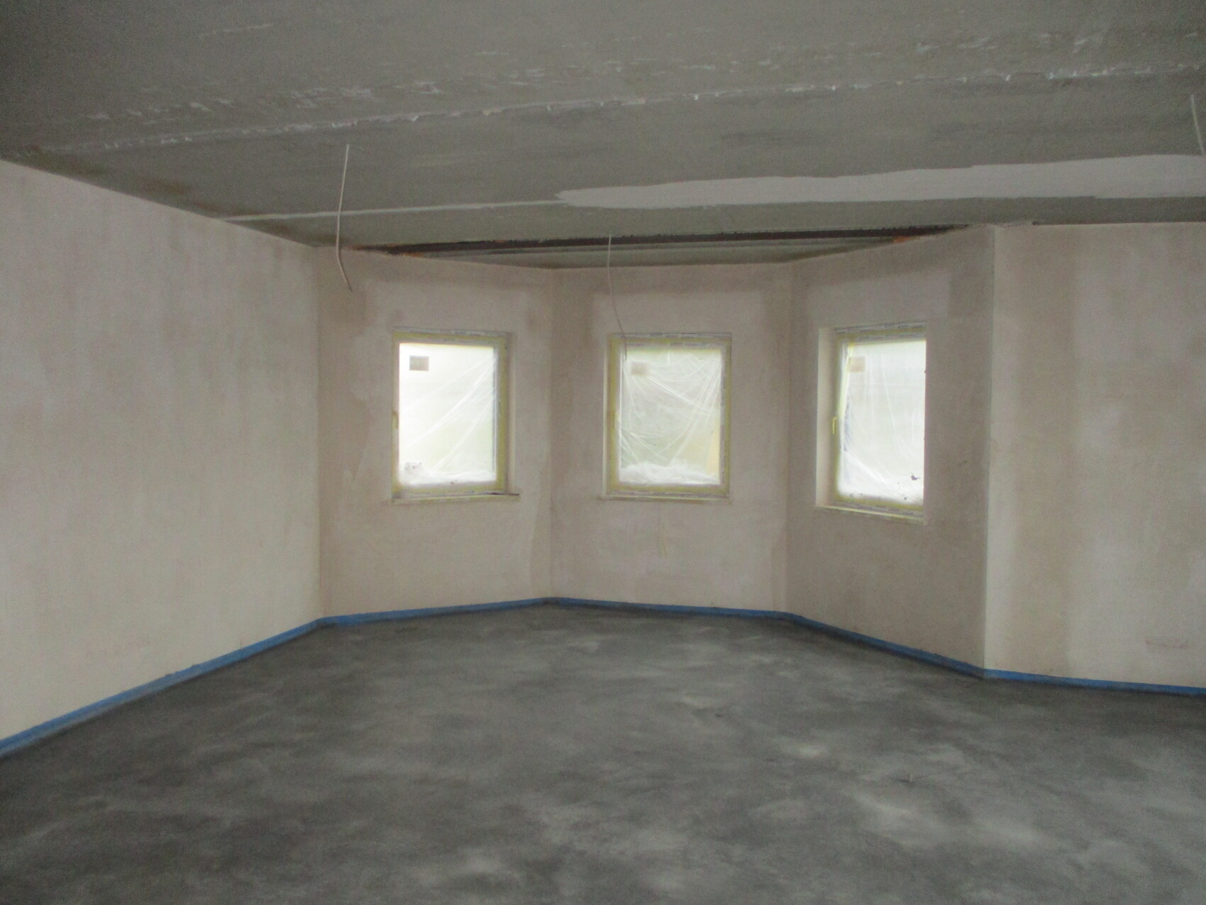 Unfertiger Innenraum: drei Fenster, kahle Wände, Betonboden, lose Kabel an der Decke.