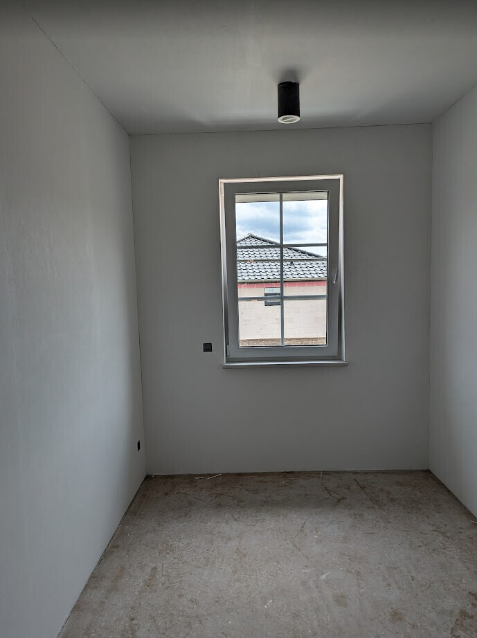 Kleines, leeres Zimmer mit grauen Wänden, Betonboden und Fenster mit Blick nach draußen.