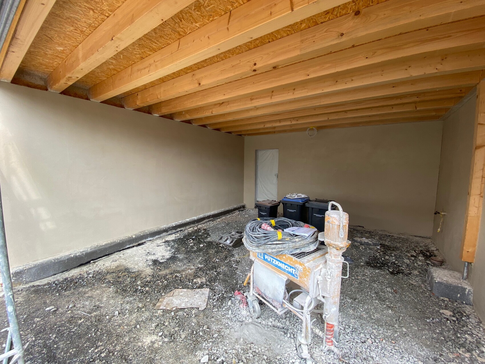 Raum auf Baustelle: Holzbalkendecke, verputzte Wände, Tür, Putzmaschine und Mülltonnen.