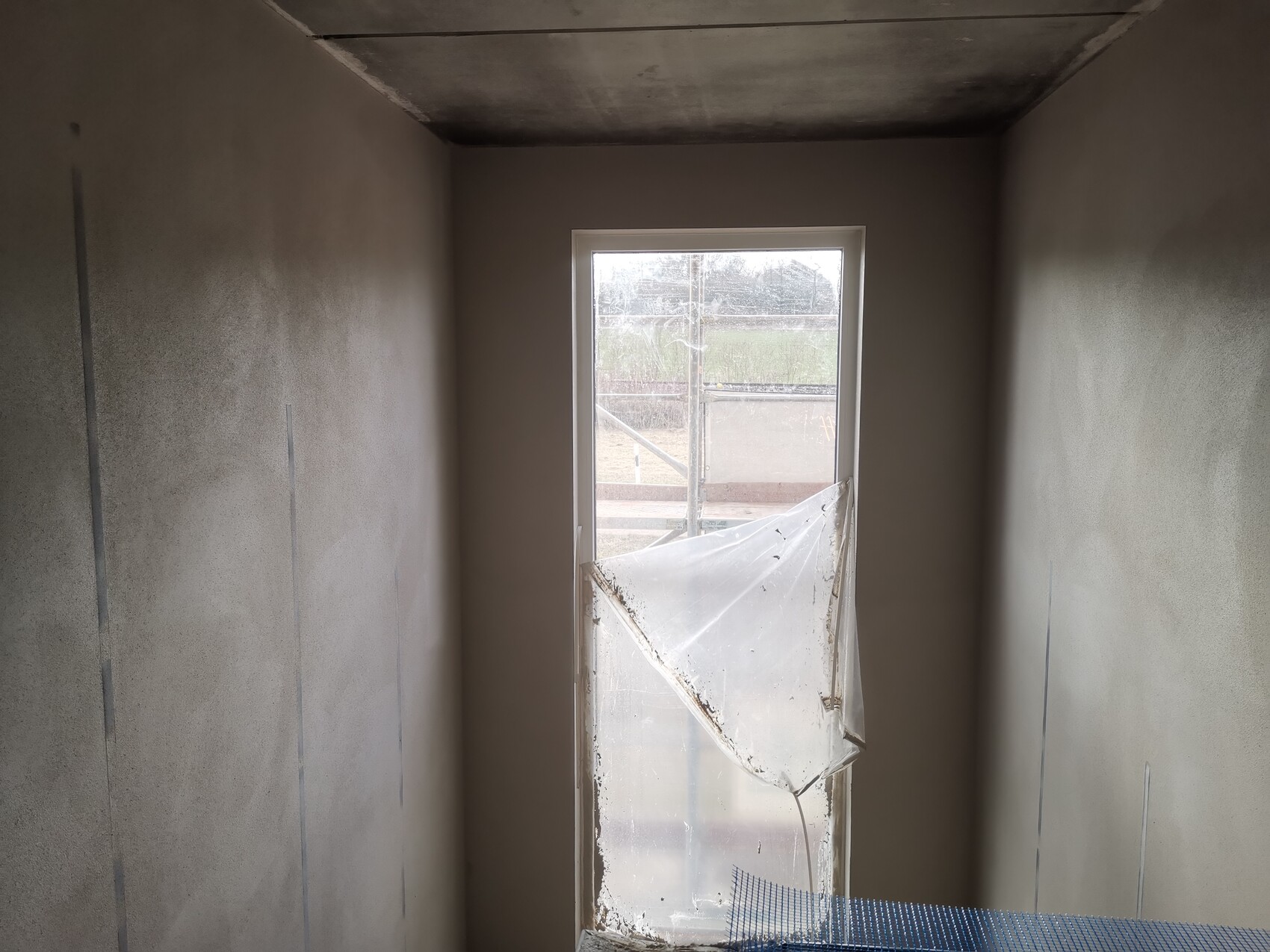 Innenraum einer Baustelle mit unfertigen Wänden, großes Fenster, beschädigte Plastikabdeckung.
