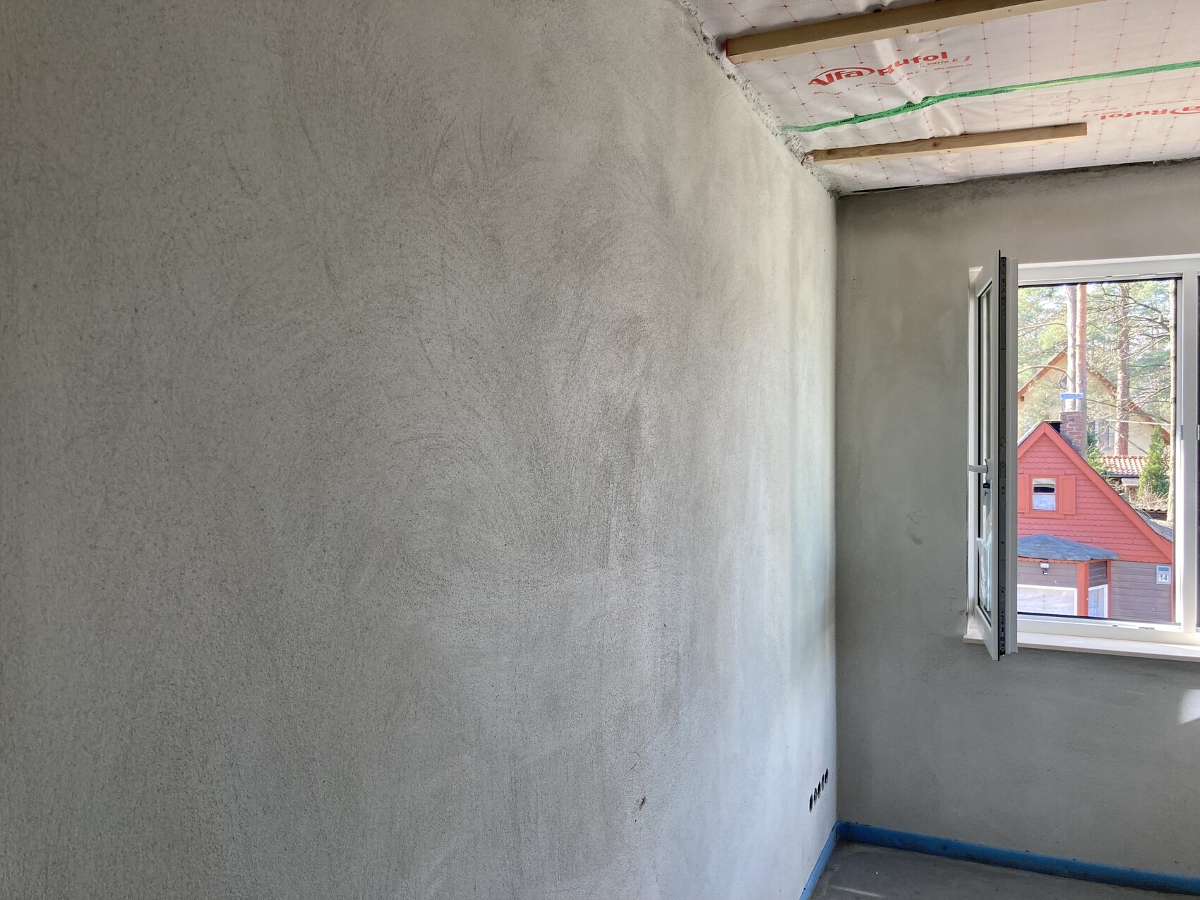 Unbearbeiteter Renovierungsraum mit grauer Wand, geöffnetem Fenster und Blick auf rotes Haus draußen.