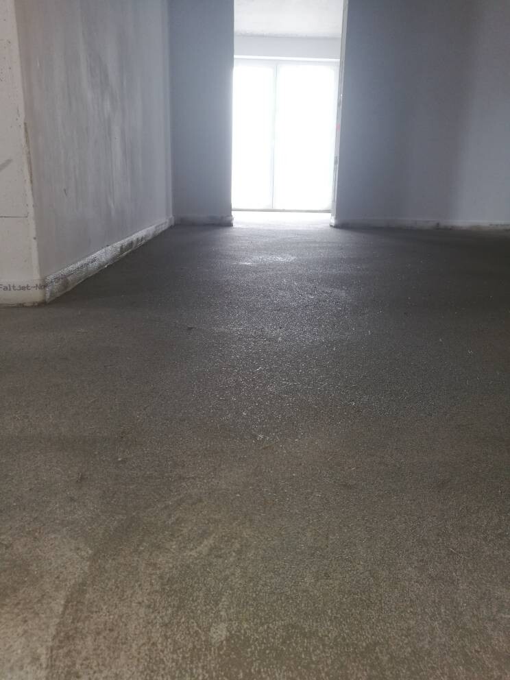 Leerer Flur auf einer Baustelle: roher Betonboden, unfertige Wände; helles Licht am Ende.