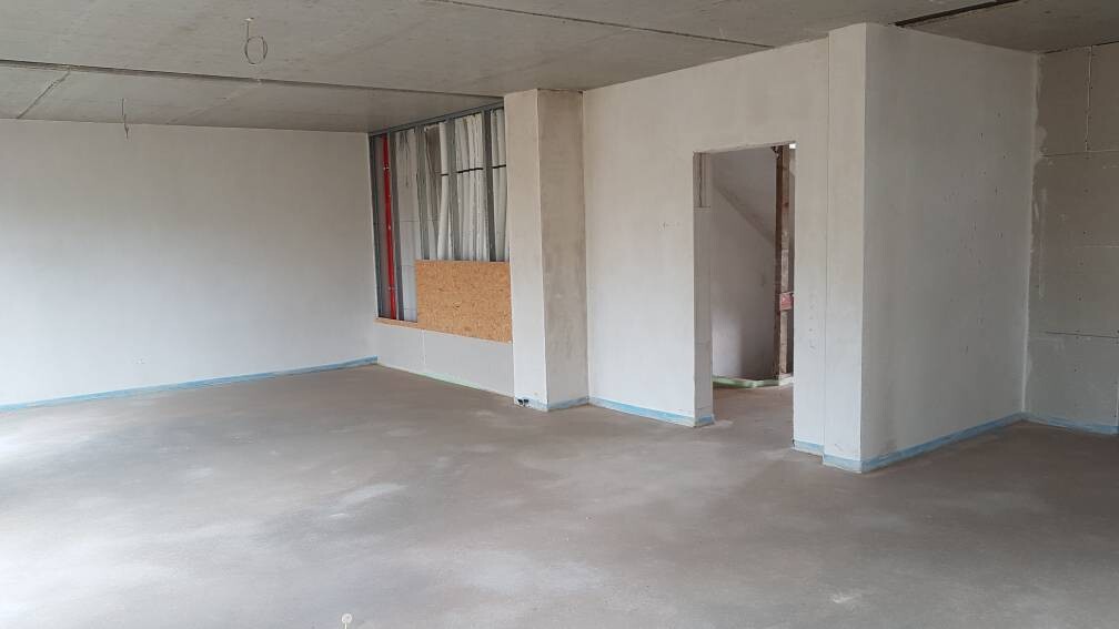 Unfertiger Innenraum einer Baustelle: Betonboden, leere Wände, Fenster mit Sperrholz, Türöffnung.