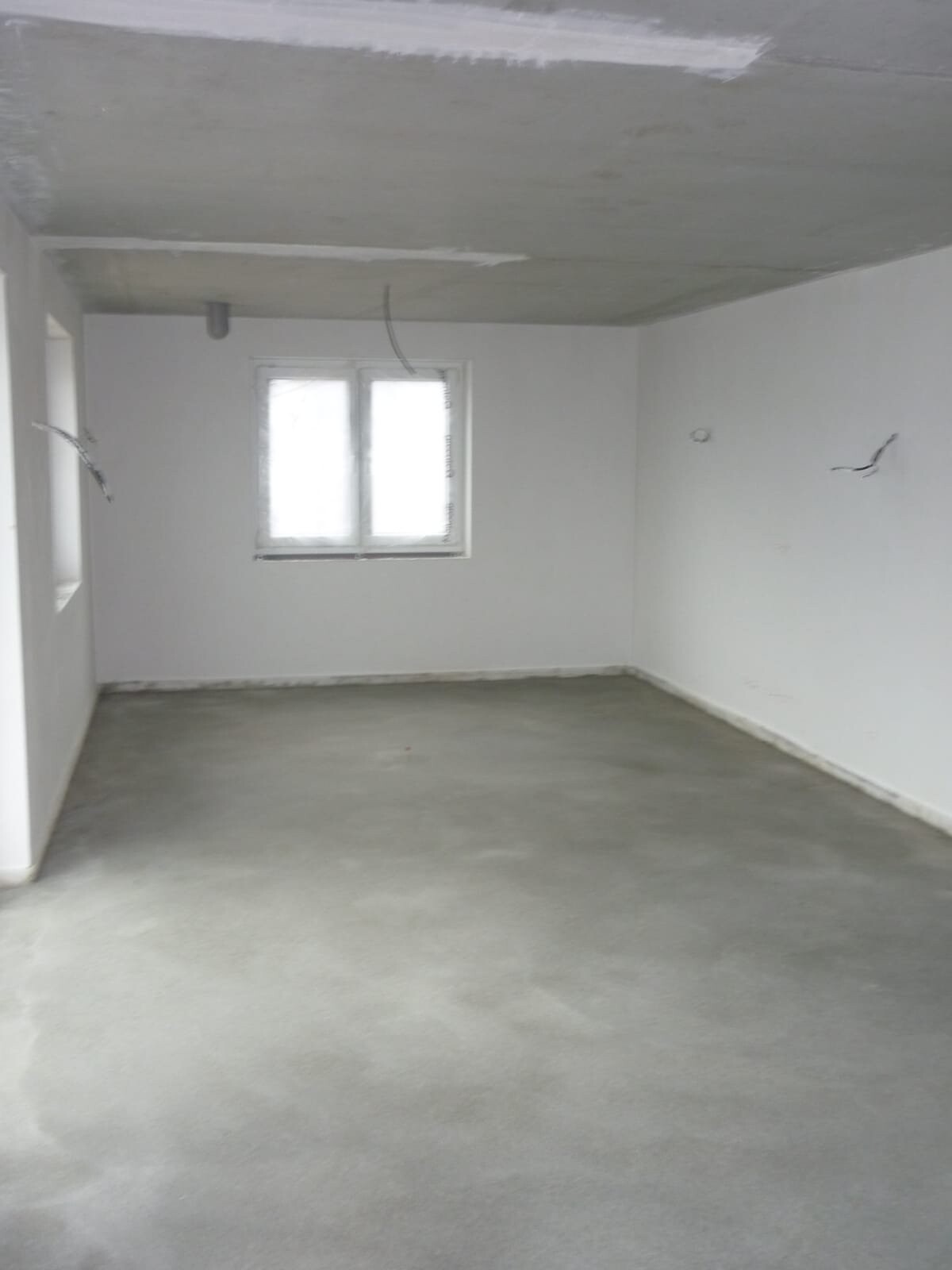 Unmöblierter Renovierungsraum mit Fenster, Betonboden, weißen Wänden und freiliegenden Kabeln.