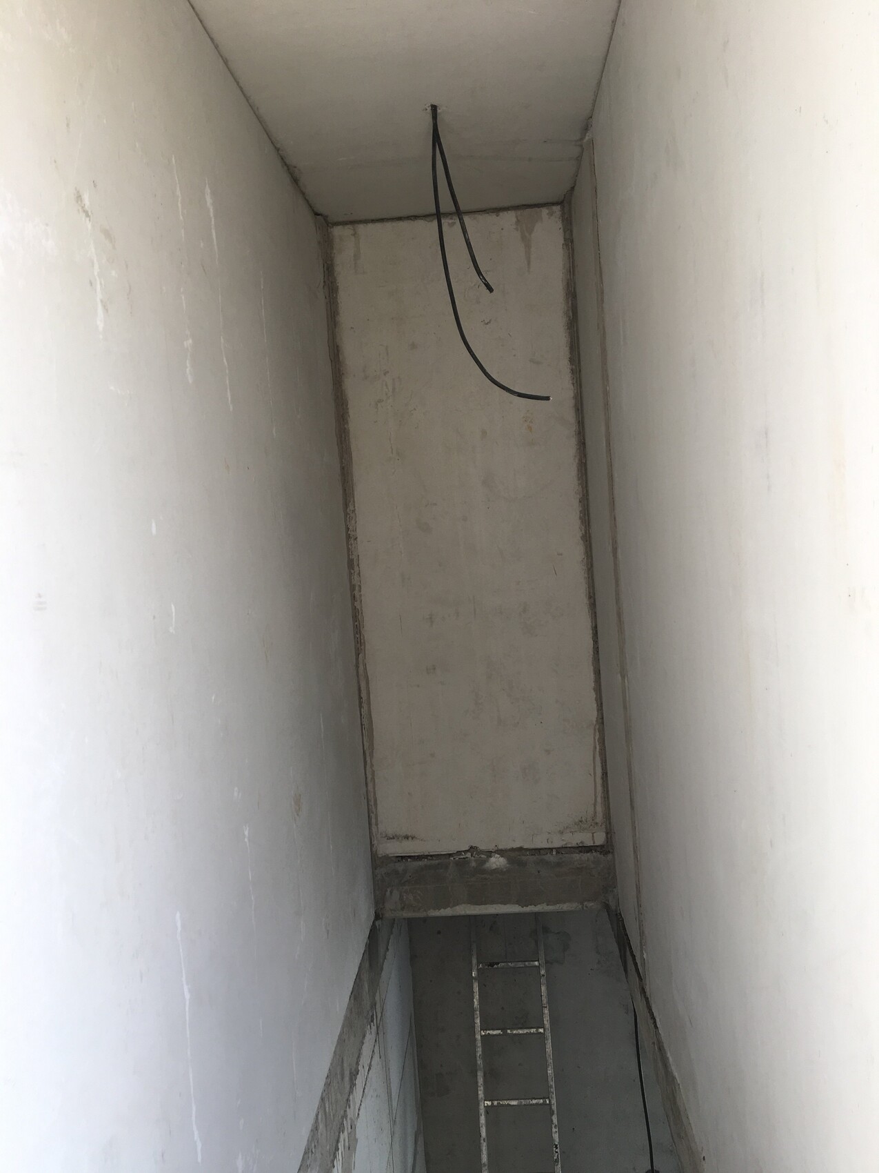 Enge Betonschachtinnenraum mit Metallleiter am Boden; Kabeln hängen von Decke herab.