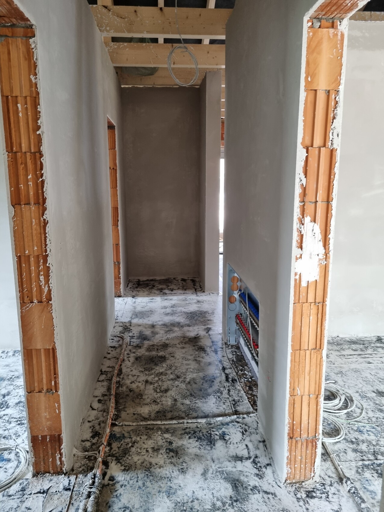 Rohbau-Gang mit Ziegelwänden, hängenden Kabeln, Holzdecke und staubigem Boden.