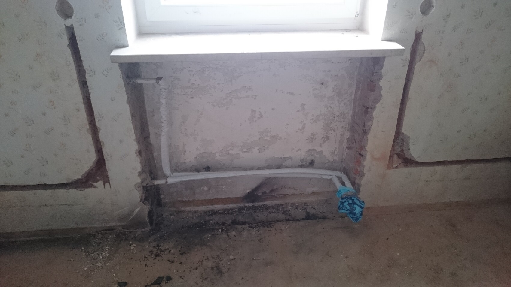 Fensterbereich einer renovierungsbedürftigen Wand mit freiliegendem Putz, Rohren und Feuchtigkeit.