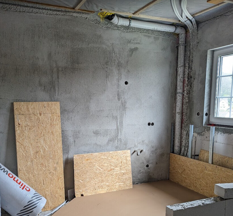 Unfertiger Kellerraum mit Rohbetonwänden, Fenster, Abwasserrohr und OSB-Platten auf dem Boden