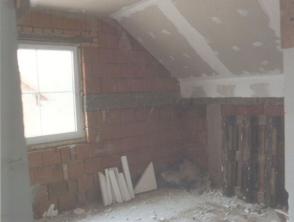 Unfertiger Innenraum mit roten Ziegelwänden, Fenster links, Bauholzreste und Trümmern.