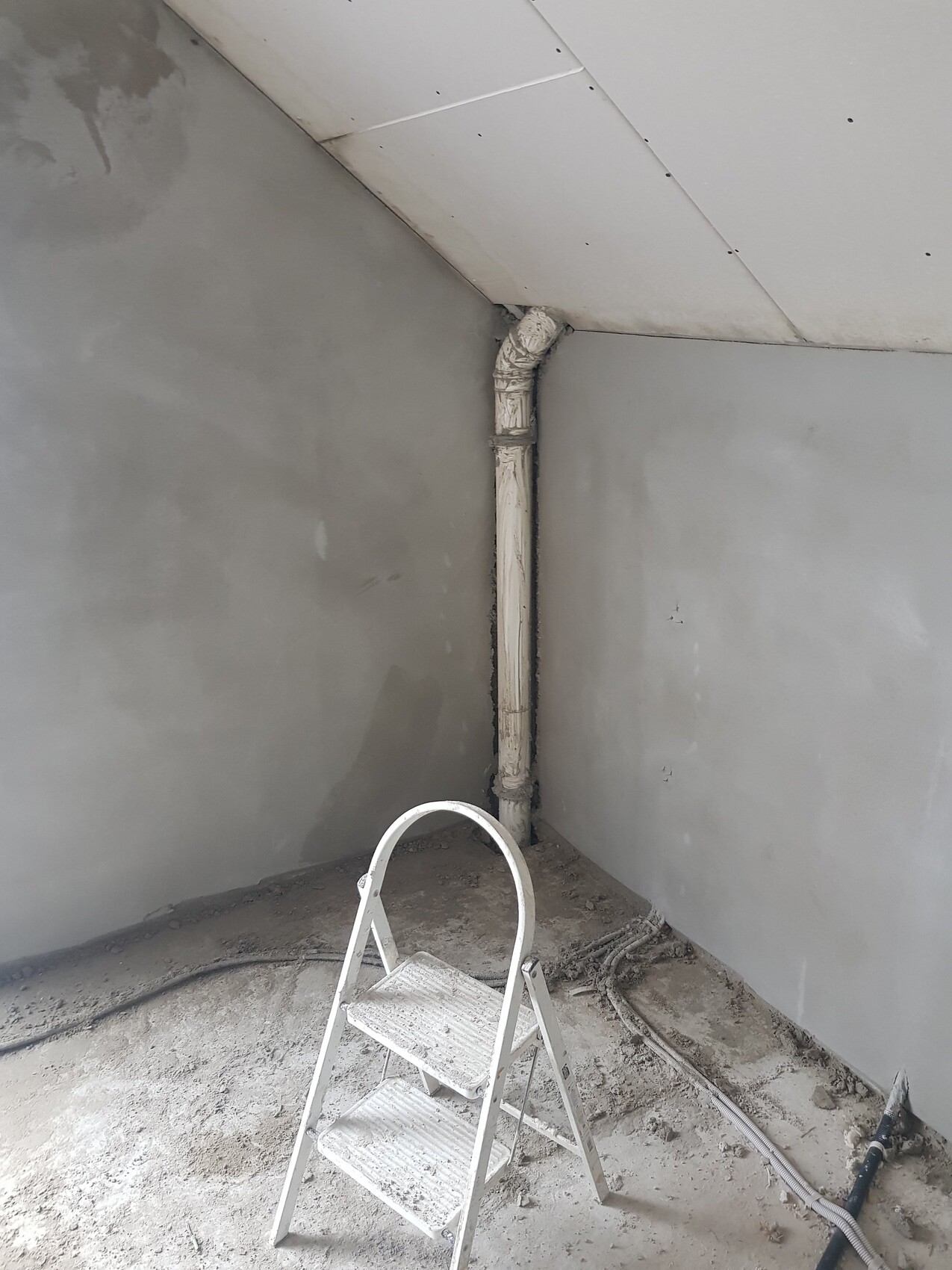 Baustellenecke: isoliertes Rohr mit Bögen an der Wand, weiße Trittleiter im staubigen Raum.