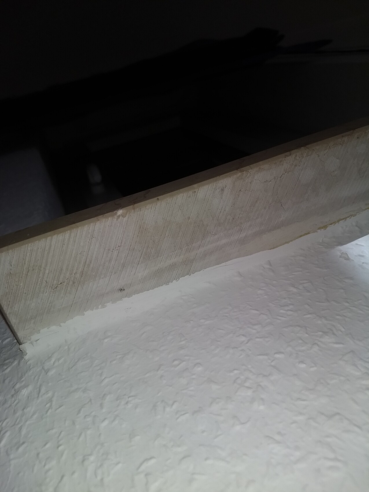 Dunkler Raum hinter einer Holzleiste; davor eine weiße, strukturierte Wand.