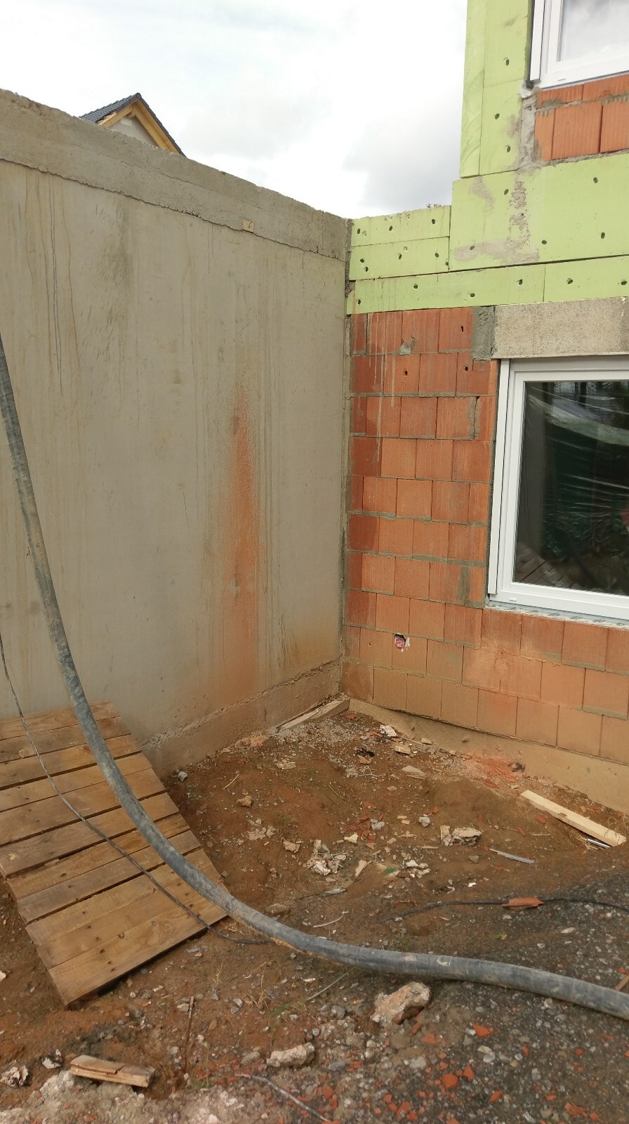 Unfertige Hauswand aus roten Ziegeln mit grünen Dämmplatten, Kabel und Bauabfall auf dem Boden.