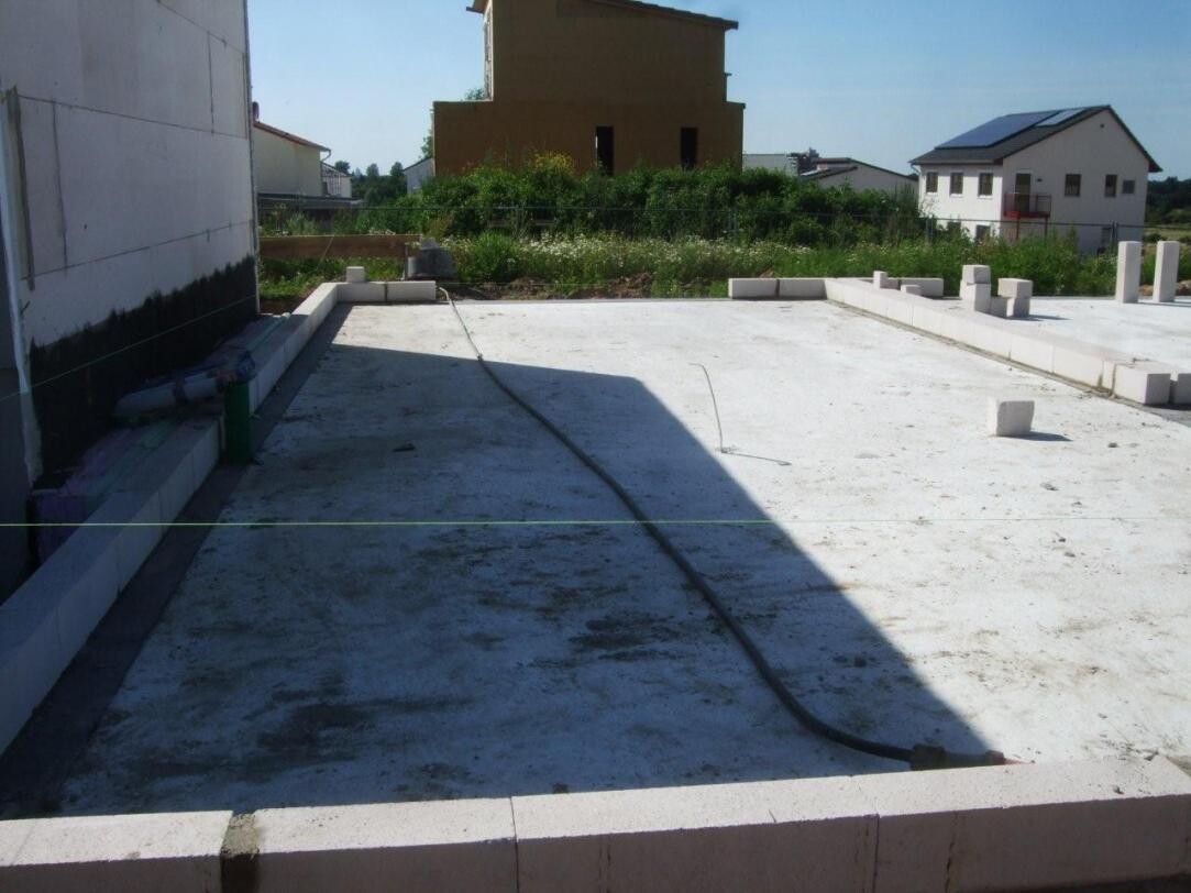 Blick auf eine Baugrube: Betonplatte, Rand aus Blöcken, Schlauch liegt darauf, Hintergrund Häuser.
