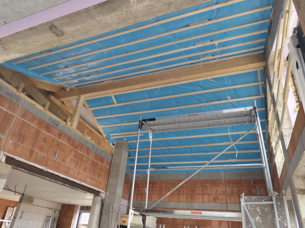 Baustelle im Innenraum: Rohbau mit Ziegelwänden, Holzrahmen, blauer Deckenisolierung und Gerüst