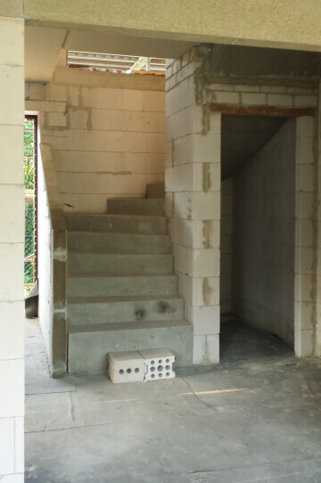 Innenaufnahme eines Rohbaus: linke Betontreppe, rechts offenes Mauerwerk, Baukonstruktion sichtbar.