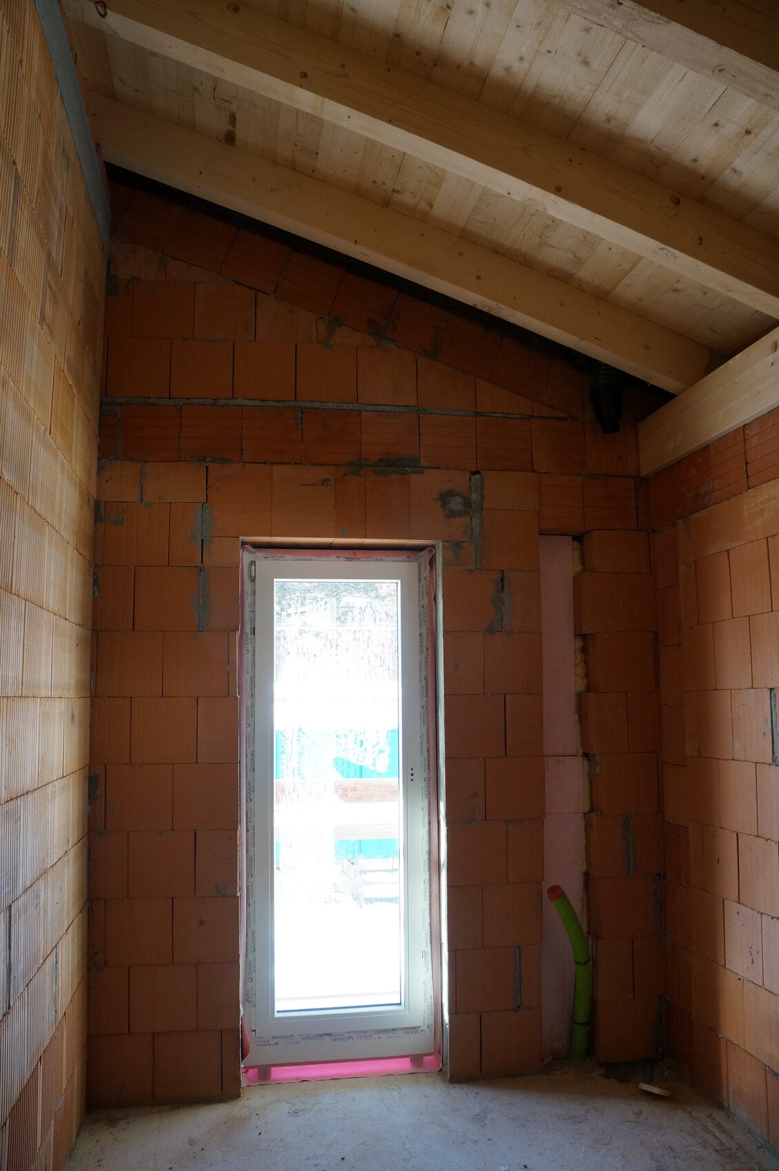 Innenraum eines Rohbaus mit roten Backsteinwänden, Holzbalkendecke und einem weißen Fensterrahmen