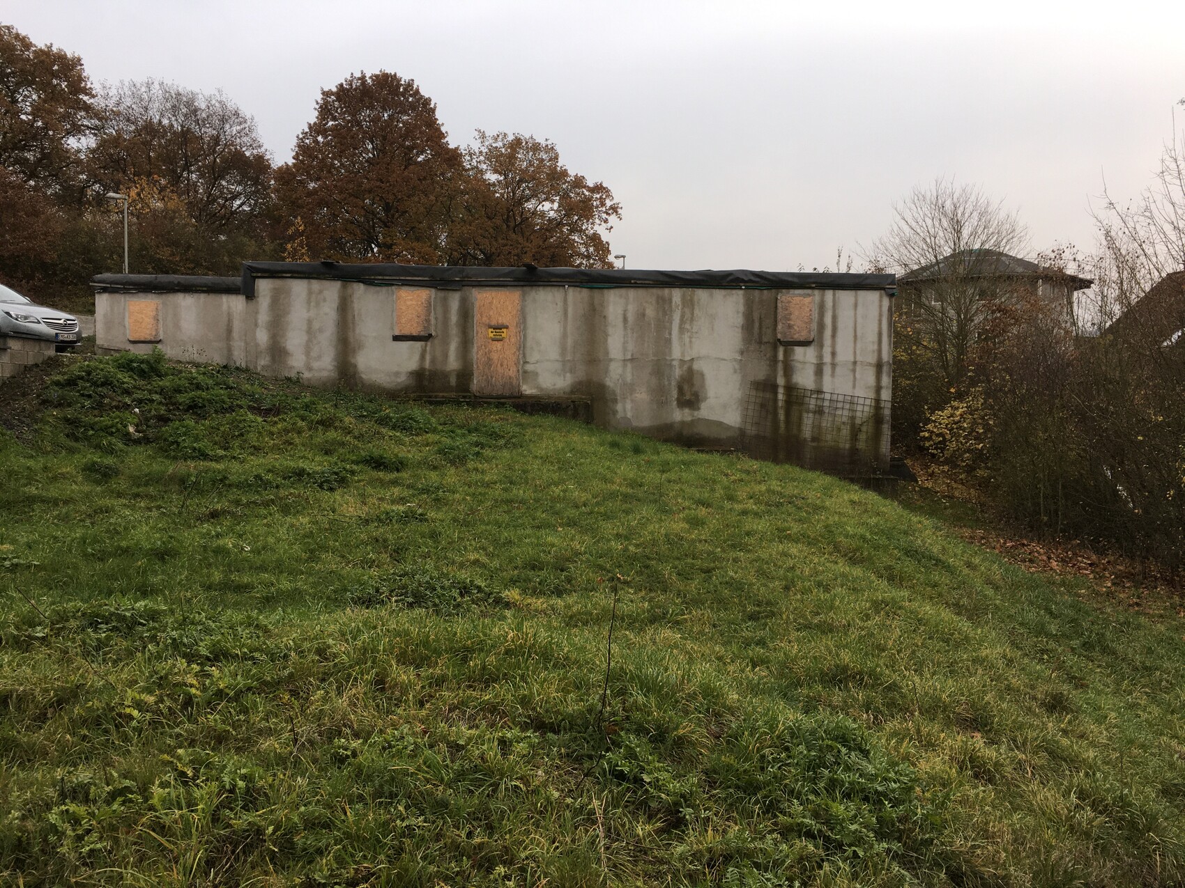 Graues Betongebäude mit Holzplatten an Türen/Fenstern, umgeben von Gras und Herbstbäumen.