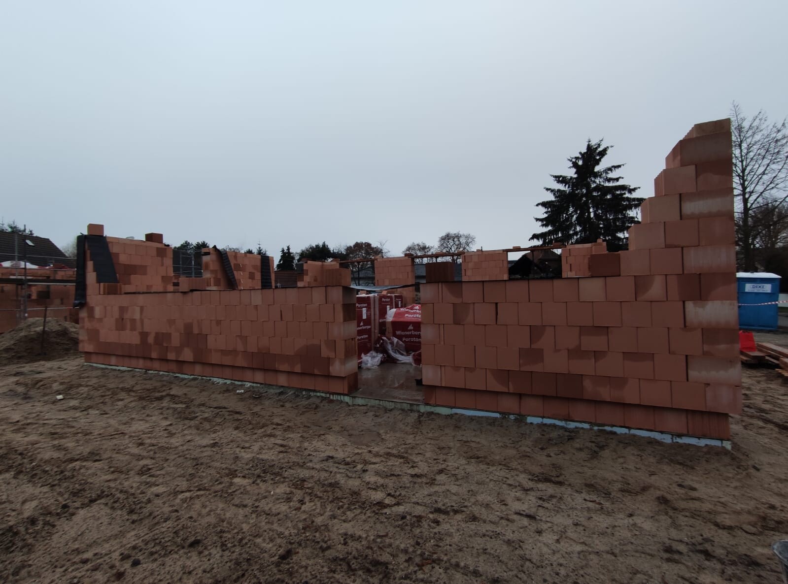 Unfertige Ziegelblockwand auf Baustelle; rote Blöcke, Sandhaufen, Bauzubehör sichtbar.