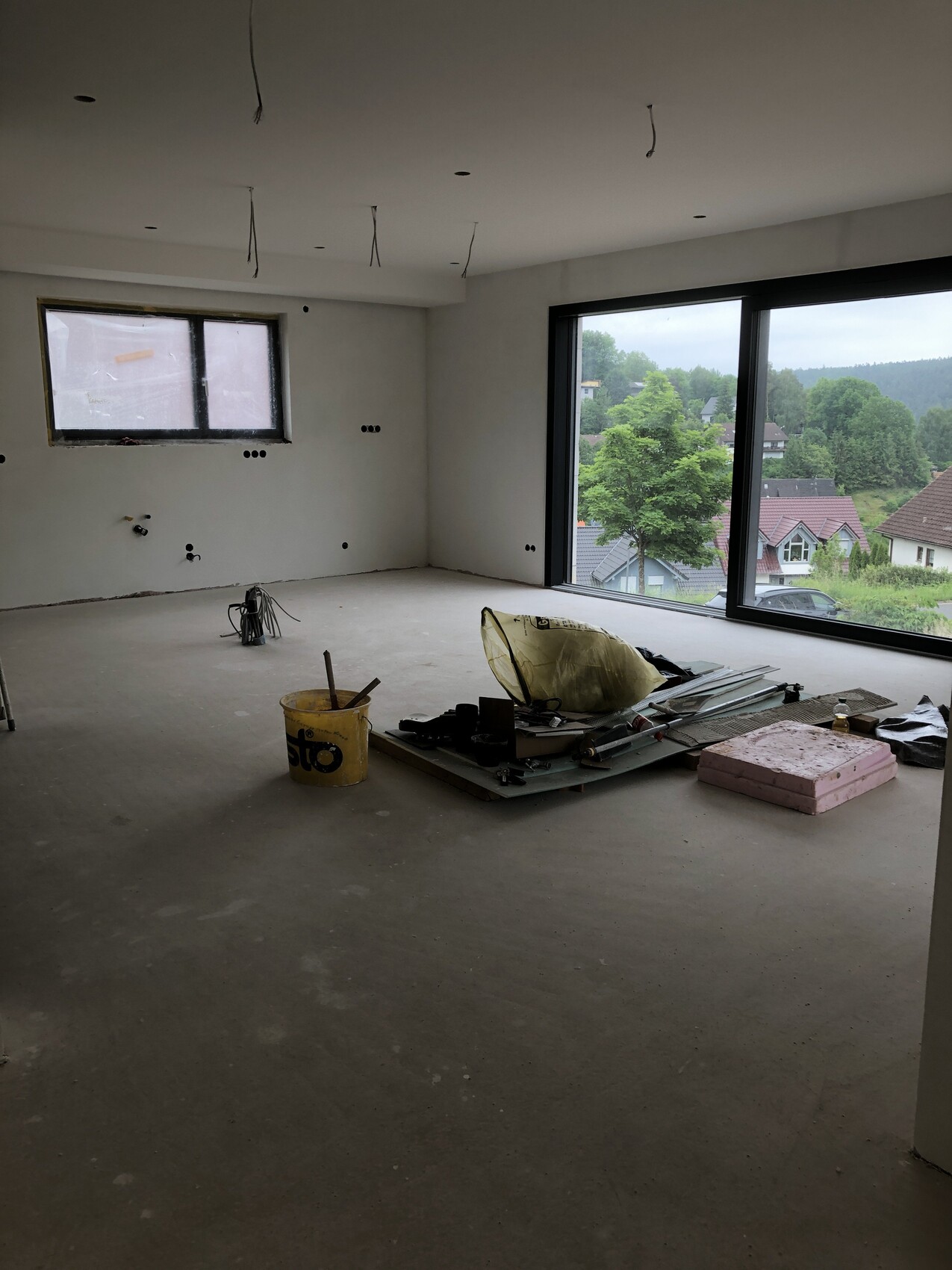 Rohbauzimmer mit Kabeln an der Decke, Bauwerkzeugen und großer Fensterfront mit Blick ins Grüne.
