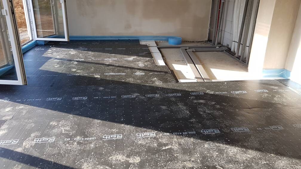 Bauzimmer mit schwarzen Unterlagsplatten, offenes Türportal und blaue Randabdichtung.
