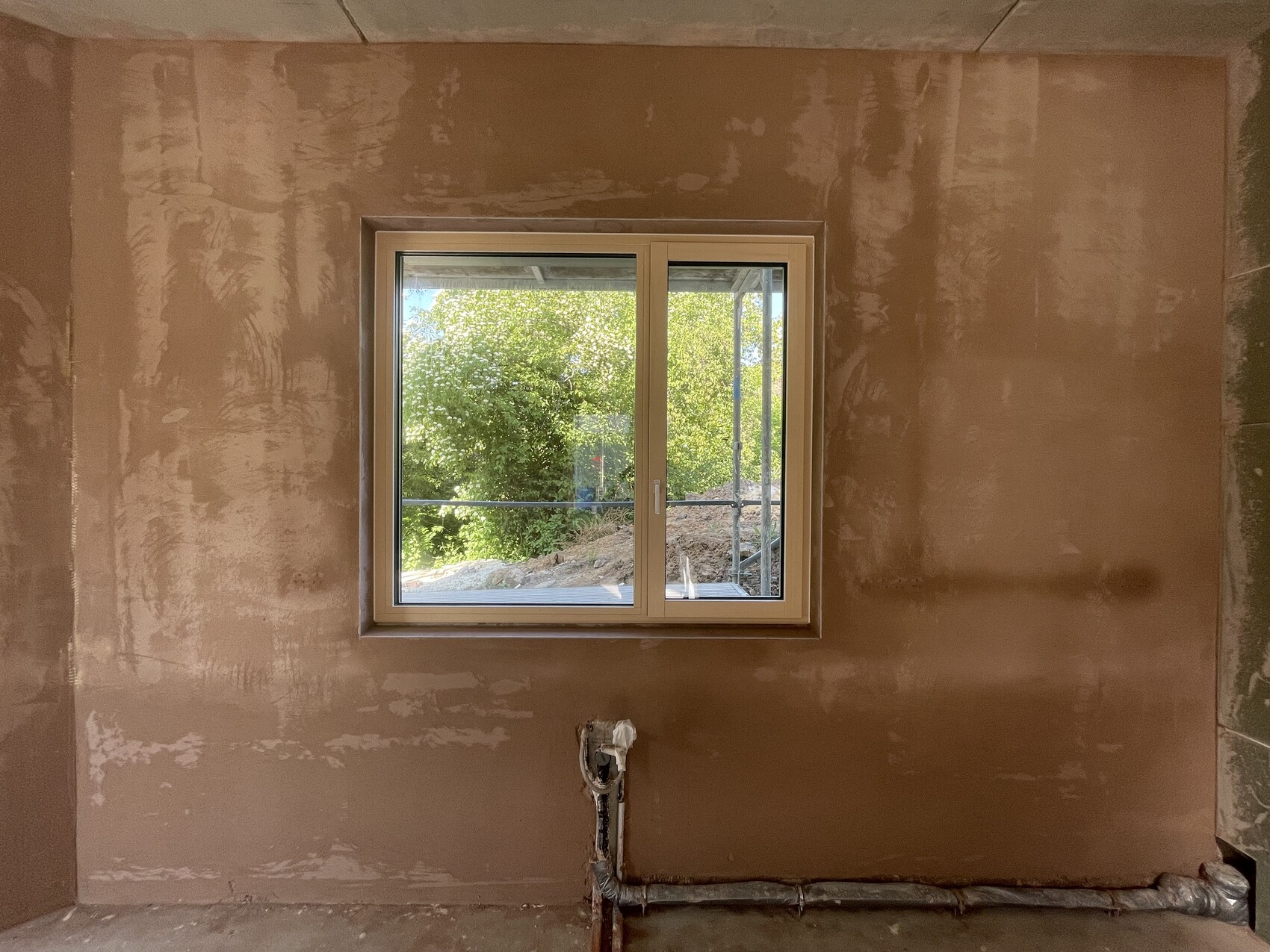 Unverputzter Innenraum mit Fenster, Blick nach außen aufs Grün, unten sichtbare Rohre.
