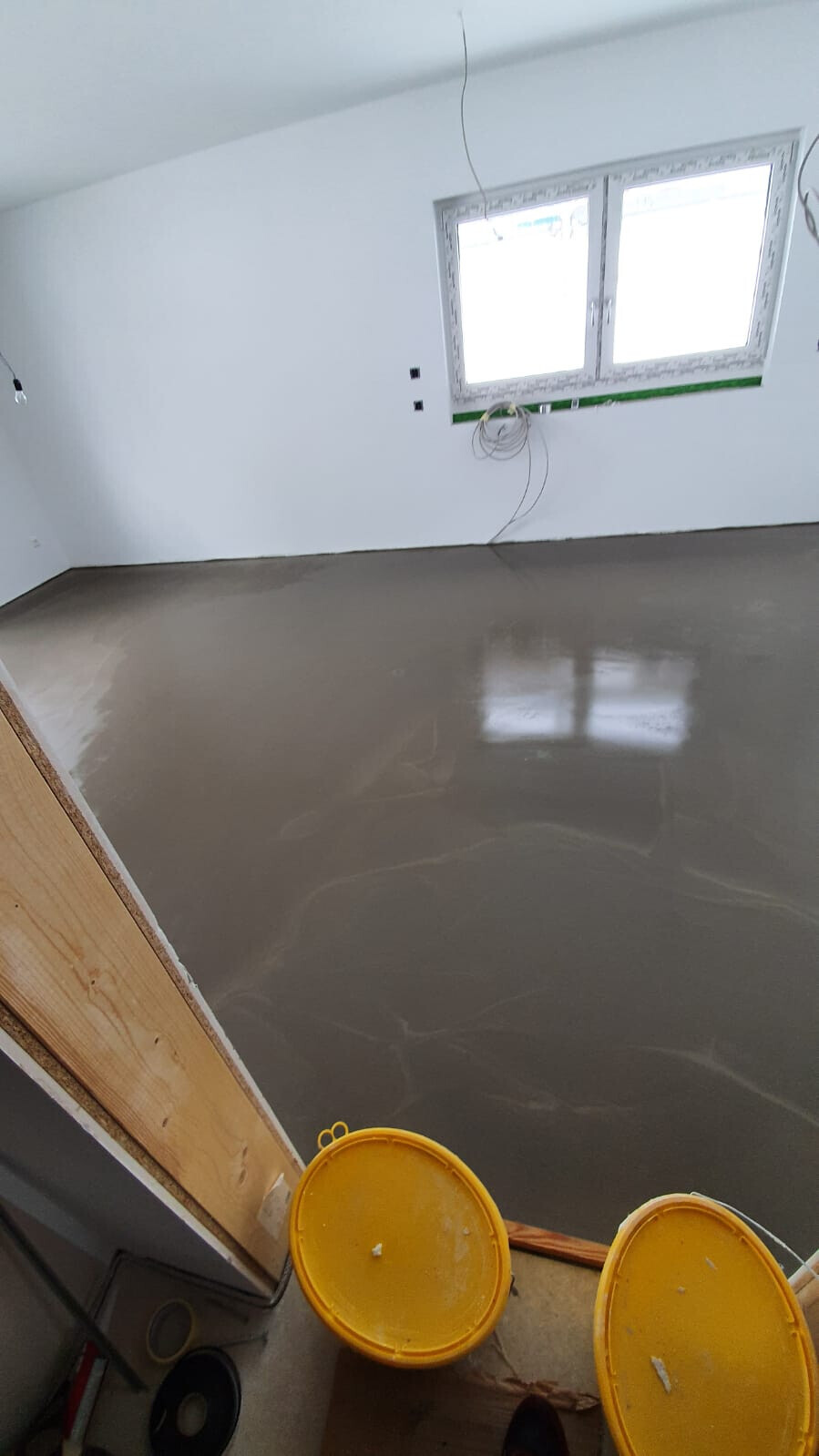 Renovierungsraum mit frisch geglättetem Betonboden, Fenster, Kabeln und zwei gelben Eimern.