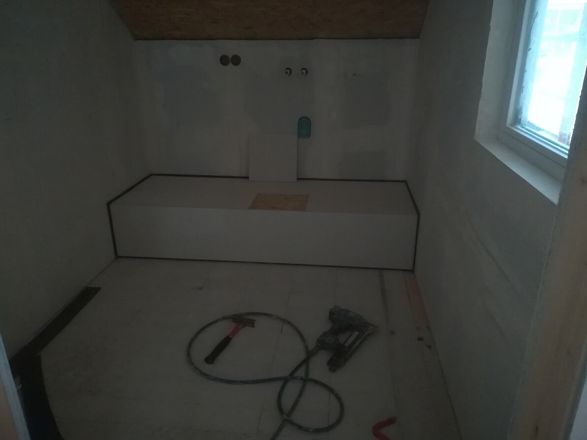 Kleine Baubaustelle: weiße Plattform gegen Wand, Werkzeuge und Kabel am Boden, Fenster rechts.
