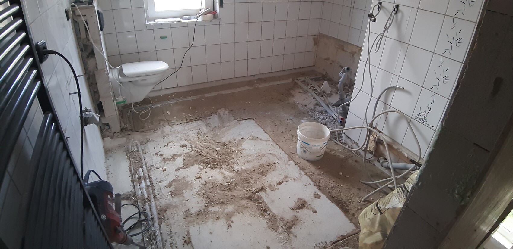 Badezimmer im Umbau: offener Boden, Schutt, Rohre sichtbar, Toilette montiert, Kabel liegen.