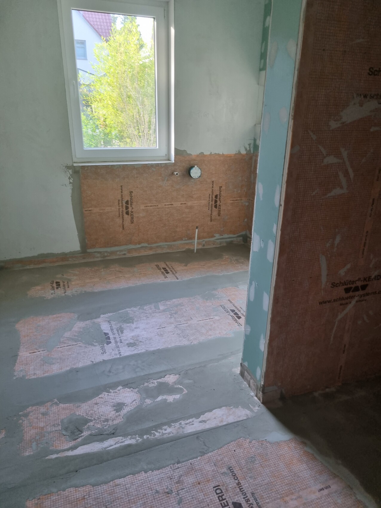 Badezimmer im Bau: Fenster, orange Abdichtungsbahn (Schluter-KERDI) an Wand/Boden, Rohre sichtbar.