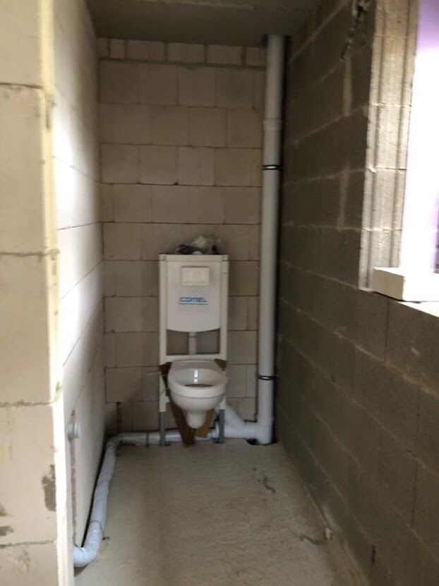 Kleines unfertiges Badezimmer mit Wandspülkasten, sichtbaren Rohren und Rohbauwänden.