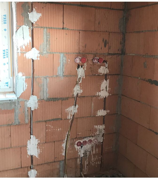 Ziegelwand mit freiliegenden Elektroanschlüssen, Kabeln und Putzresten, Fenster links.